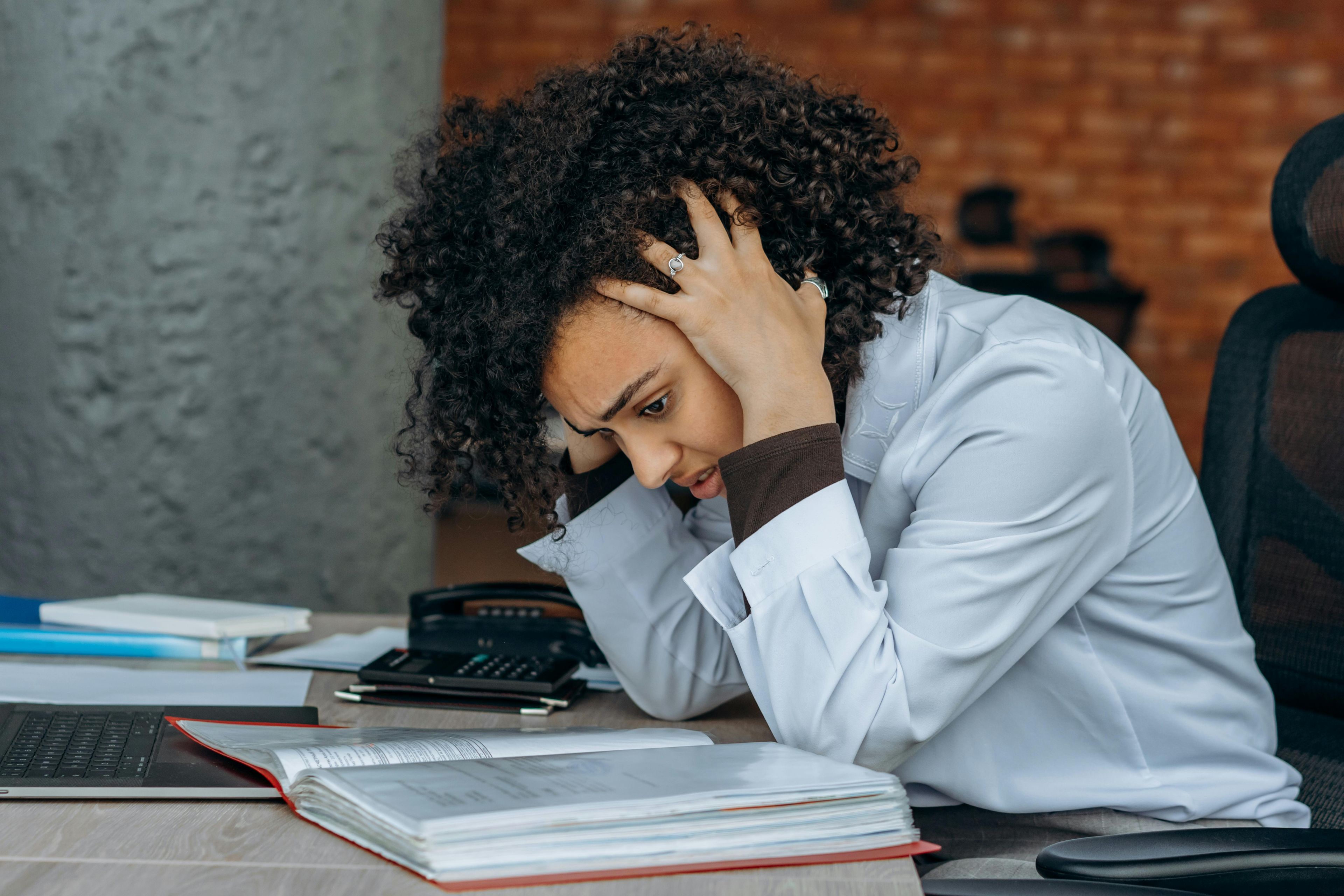 A person in a white lab coat holds their head in frustration while looking at an open binder at a desk 